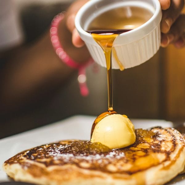 A hand pouring syrup from a small dish onto pancakes and a pat of butter